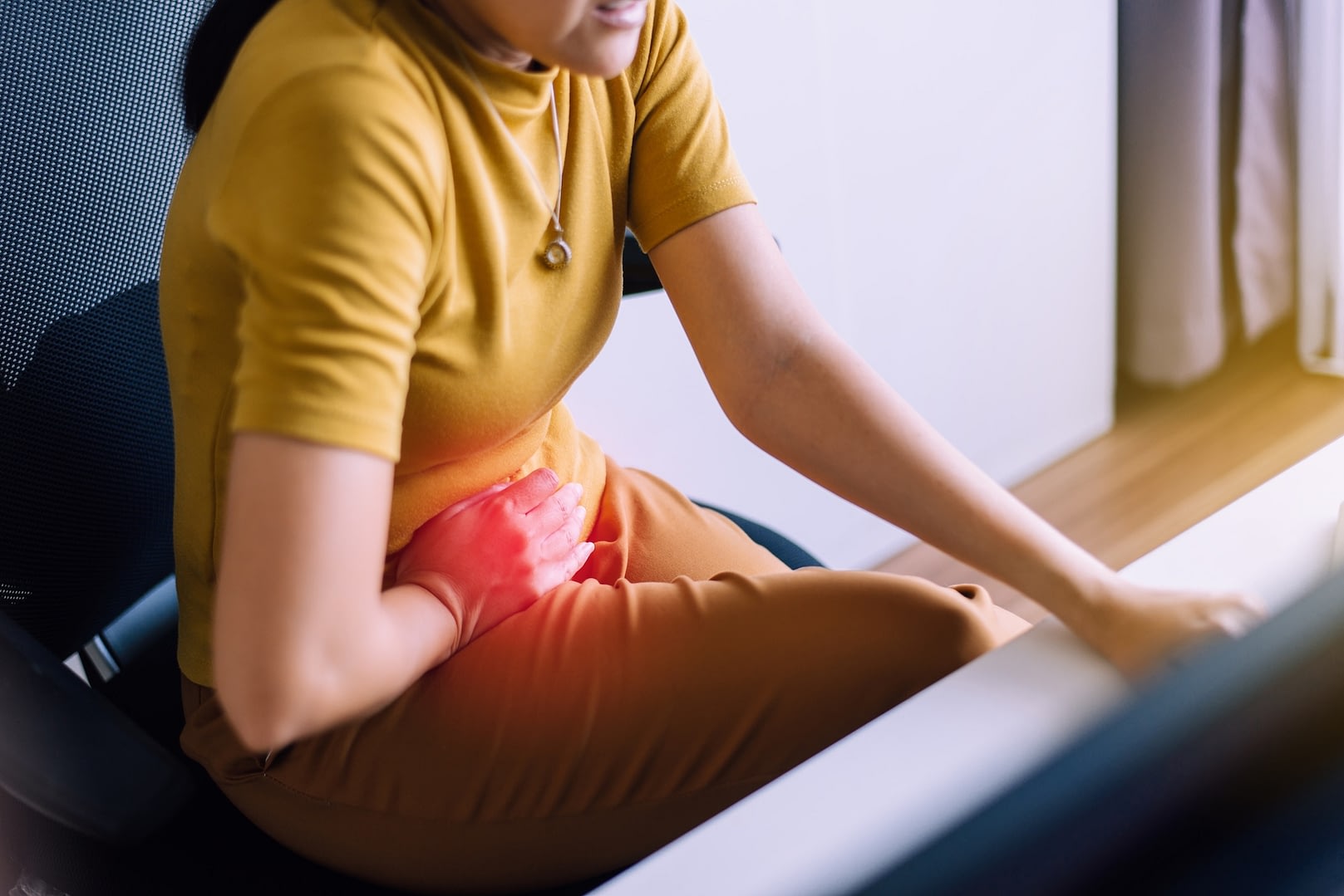 Young woman sat at desk, gripping stomach from pain.